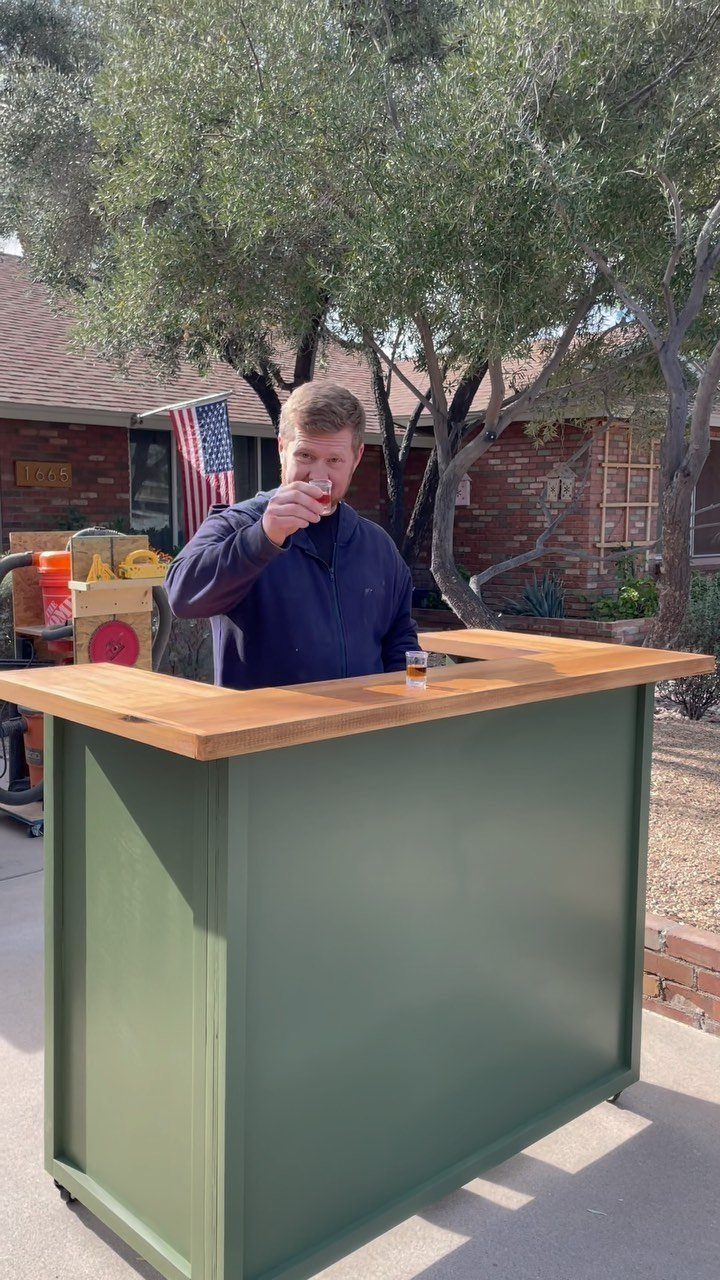 Man drinking behind a green outdoor bar with a wooden countertop on a patio