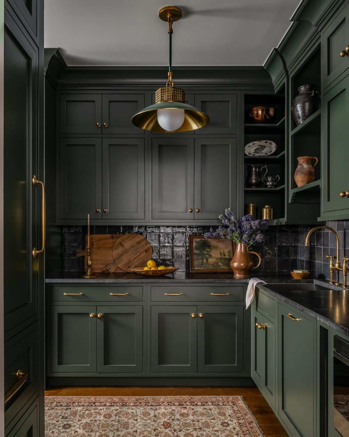 Dark green kitchen cabinets with brass hardware, black tile backsplash, and gold lighting.