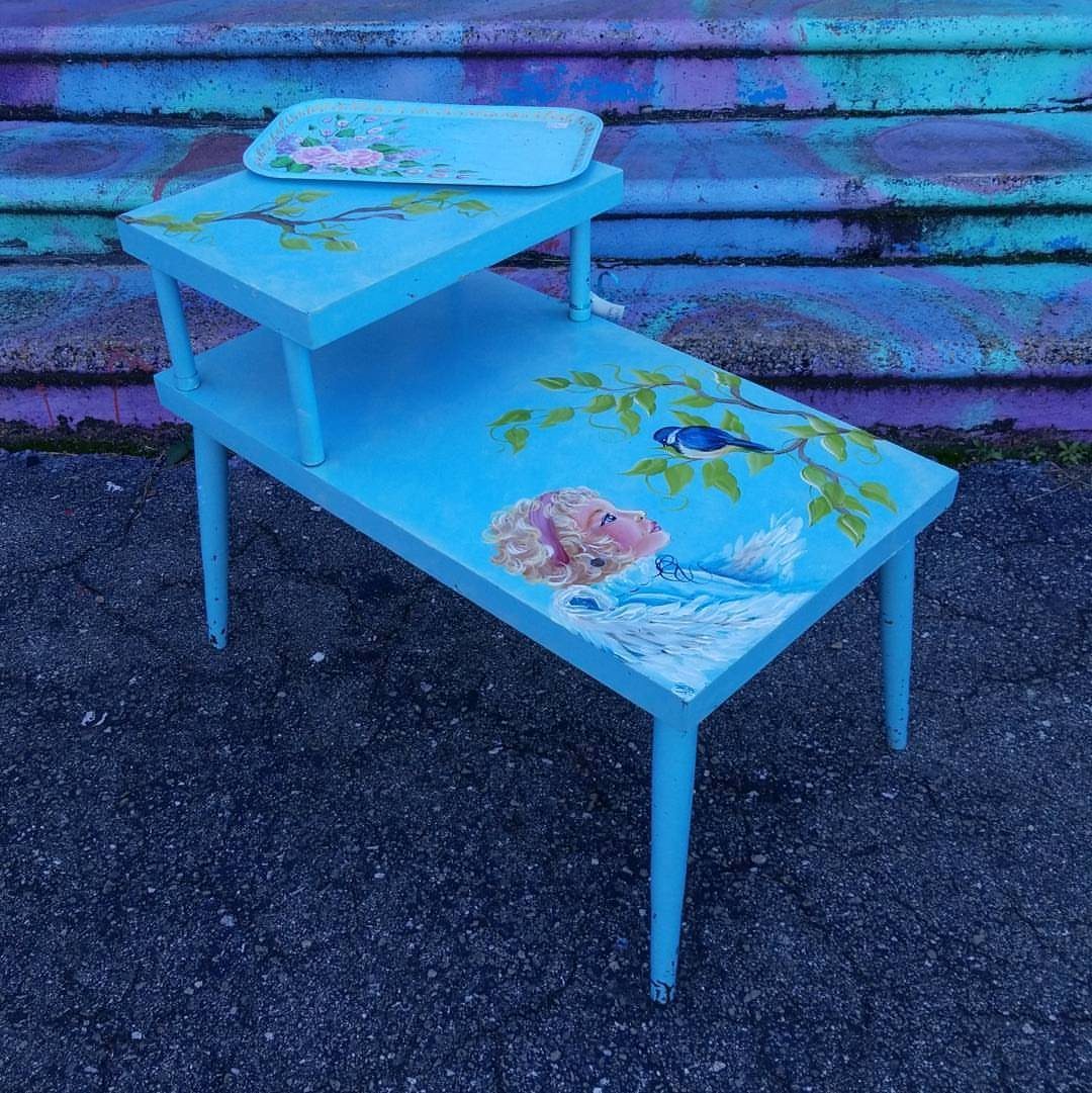 Light blue painted wooden table with cherub and bird artwork on asphalt