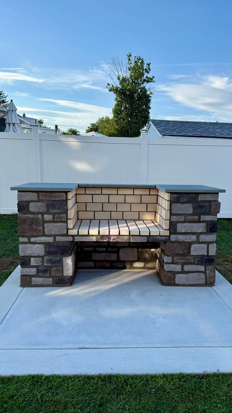 Stone and brick outdoor grill station on a concrete pad in a backyard.