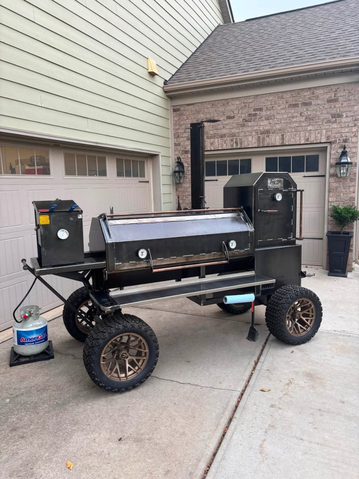 Black metal offset smoker grill on a wheeled trailer parked on a driveway.