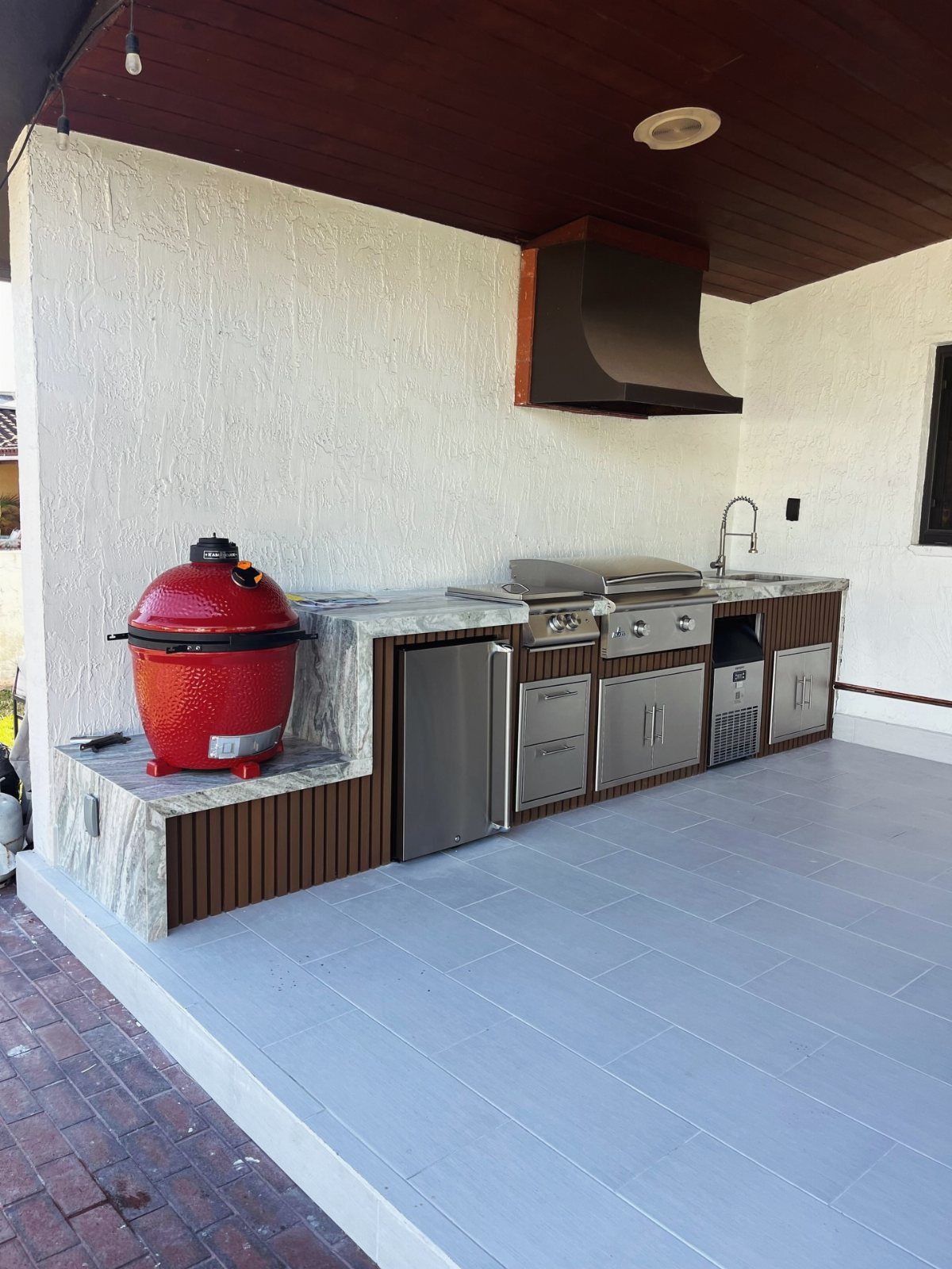 Outdoor kitchen with red ceramic grill and stainless steel appliances under wood ceiling