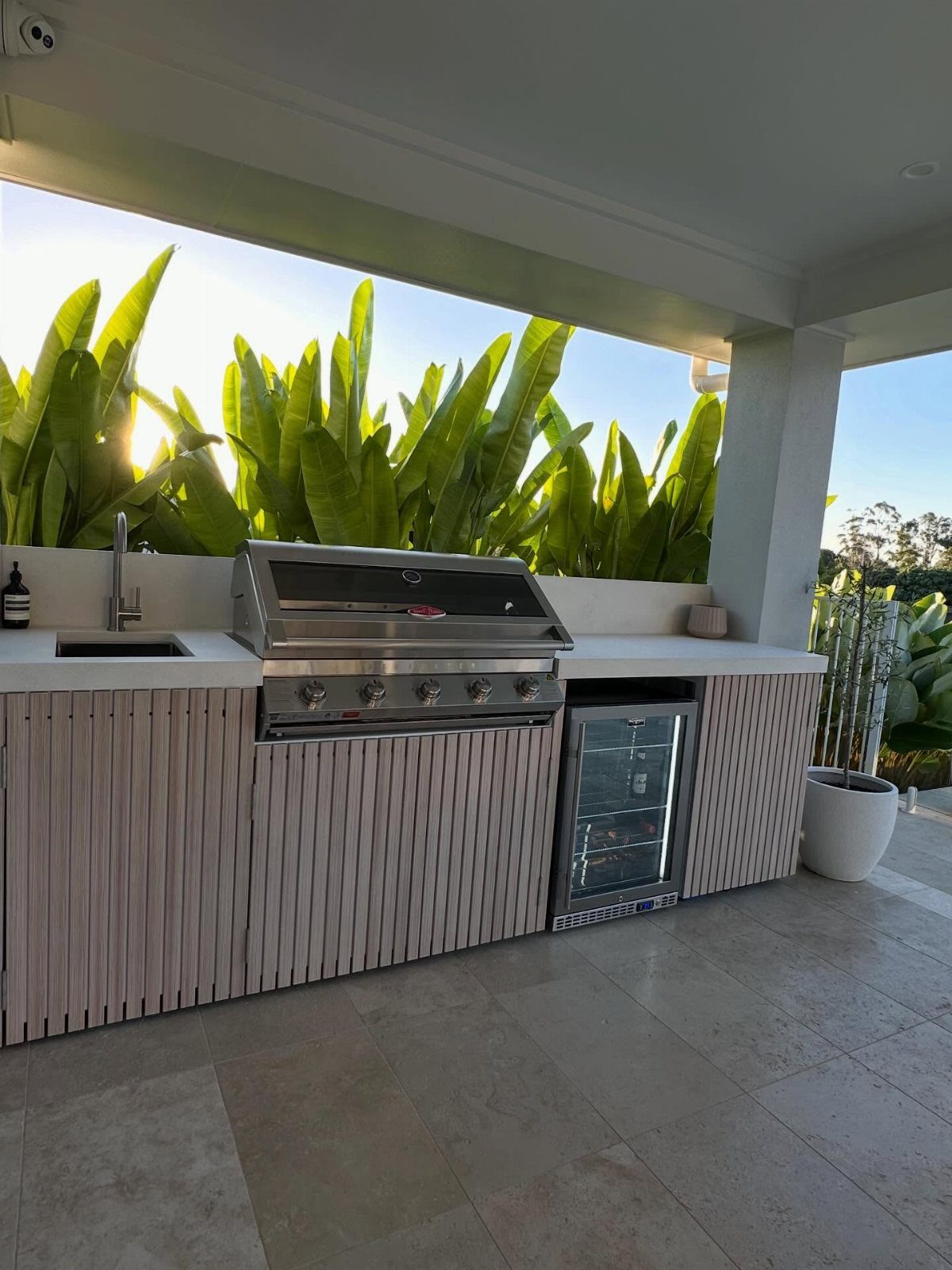 Outdoor kitchen with grill and sink under a white ceiling with tropical plants.