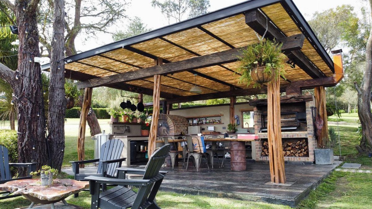 Rustic outdoor kitchen with thatched roof, brick oven, and wooden log supports.