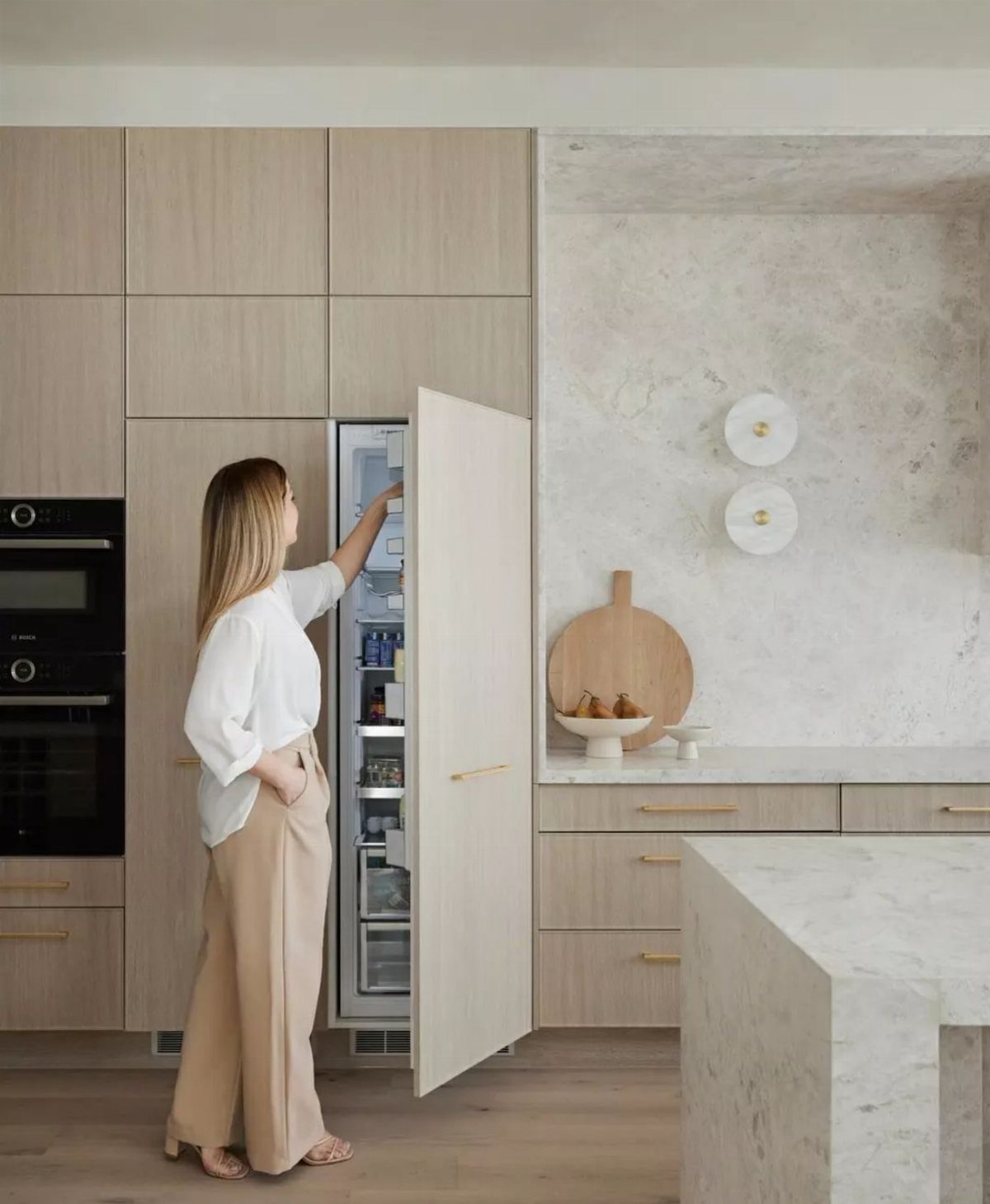 Woman opening a built-in refrigerator with matching light wood cabinetry and marble countertops.