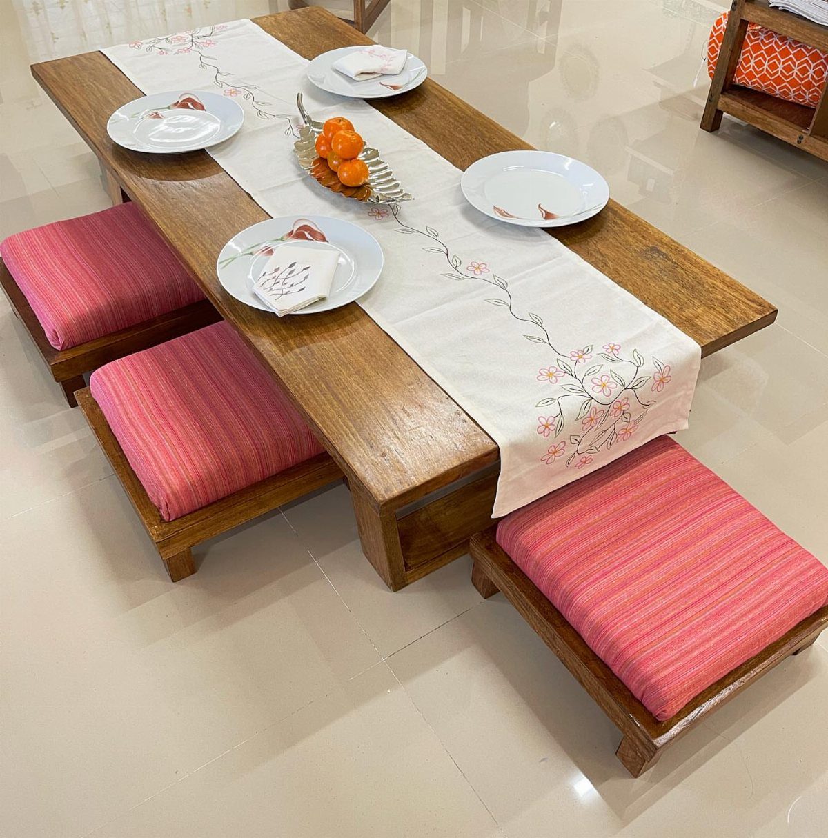 Low wooden dining table with pink striped floor cushions and white floral runner.