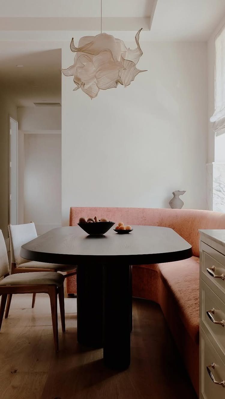 Dark oval dining table with a peach upholstered banquette and sculptural white pendant light.