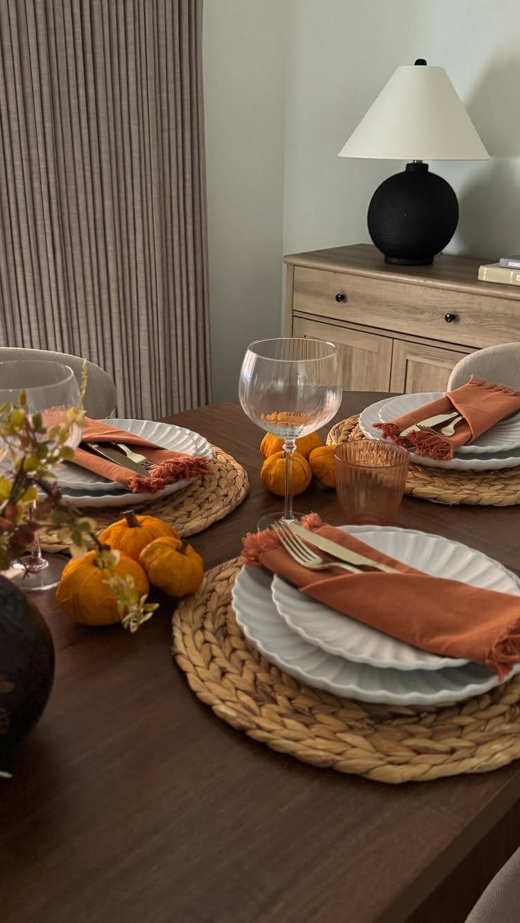Dining table set with orange napkins, woven placemats, and small decorative pumpkins.