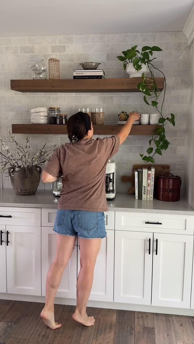 Woman reaching for items on floating wooden open shelves in a white kitchen