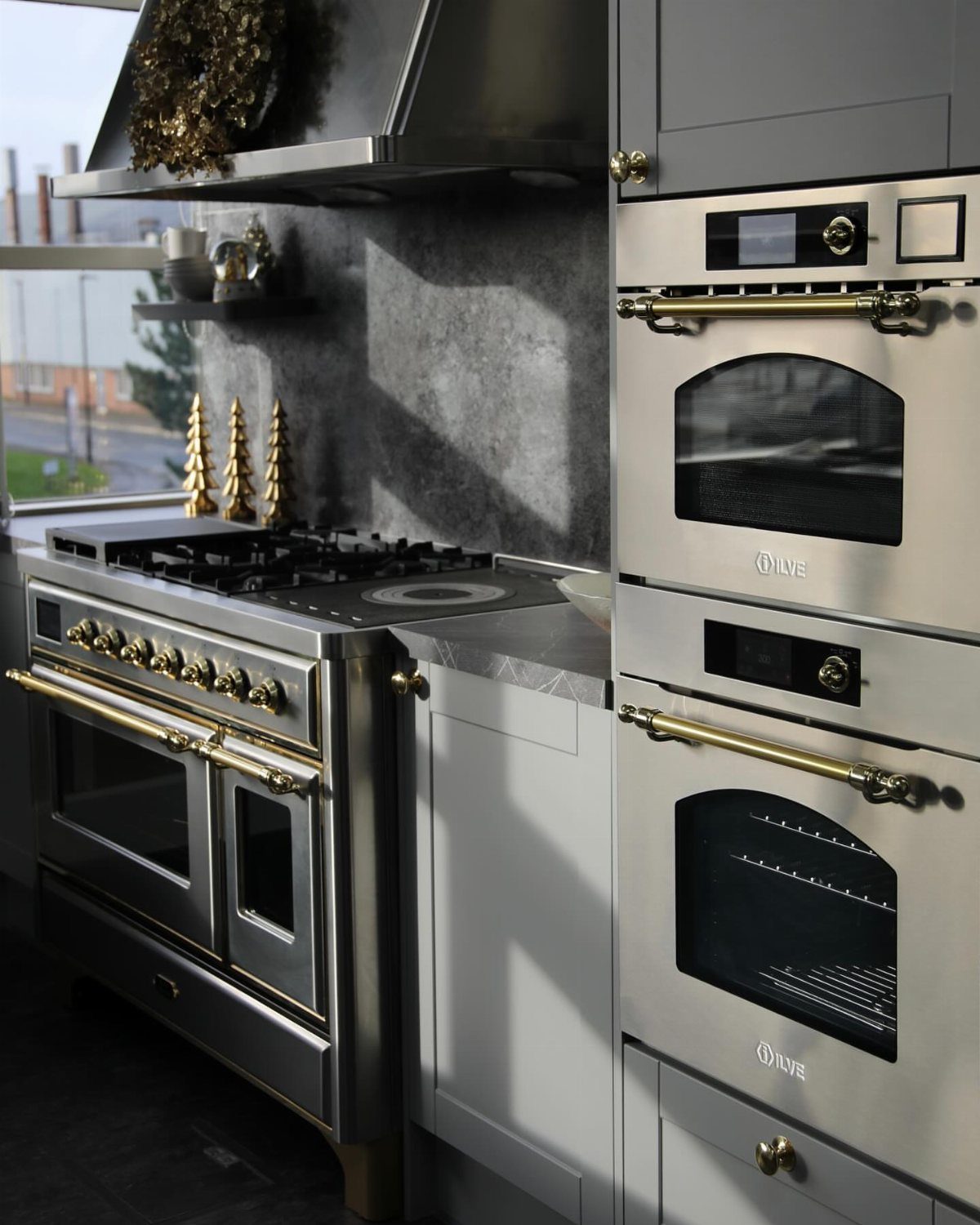 Stainless steel range and wall ovens with gold handles in a grey kitchen.
