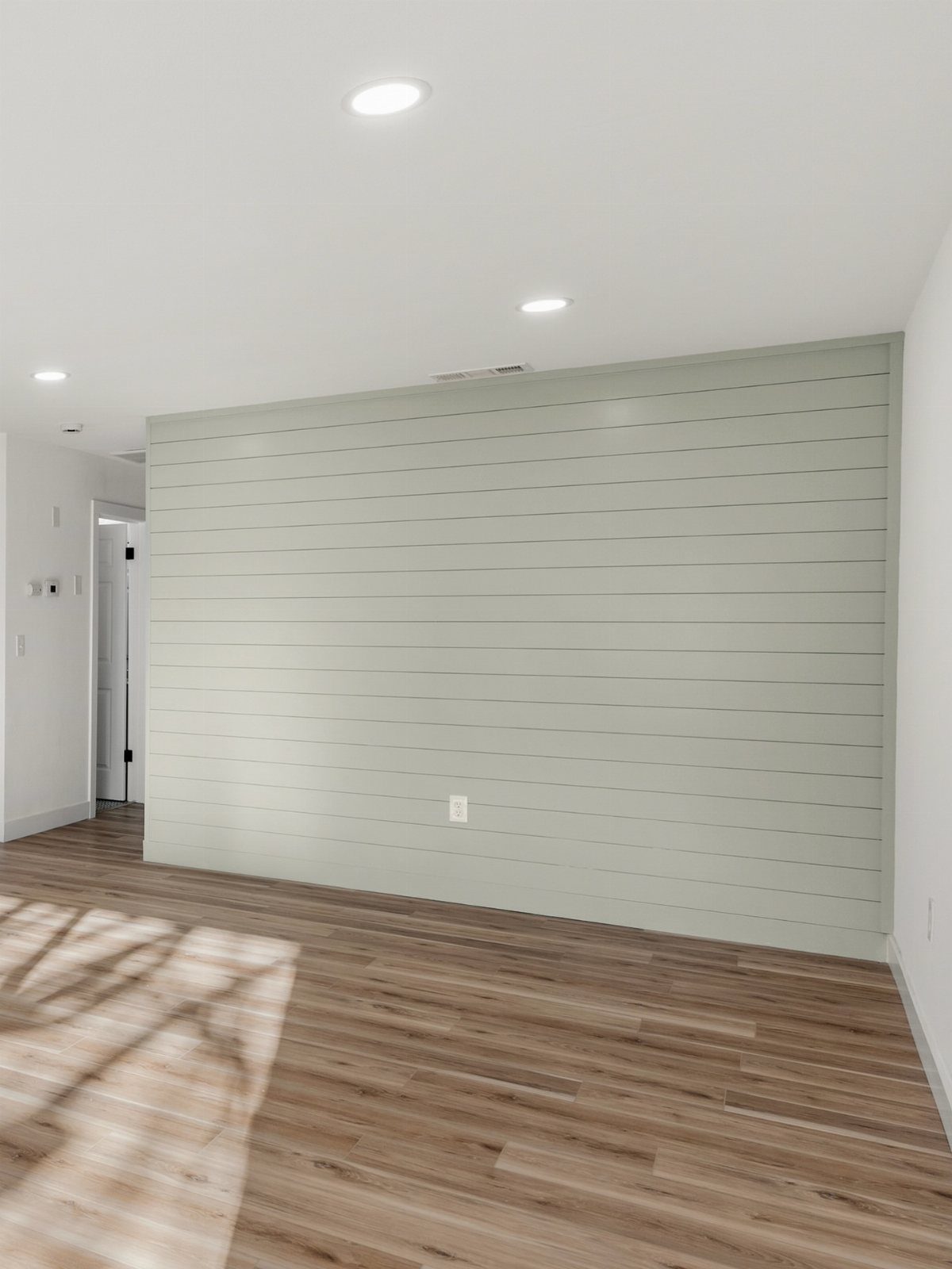 Light sage green shiplap wood paneling accent wall in a room with wood floors.
