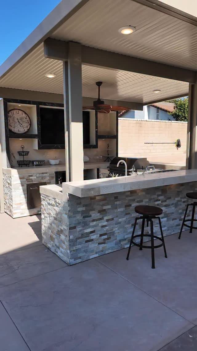 Covered outdoor kitchen with a stone bar, built-in grill, and black bar stools.