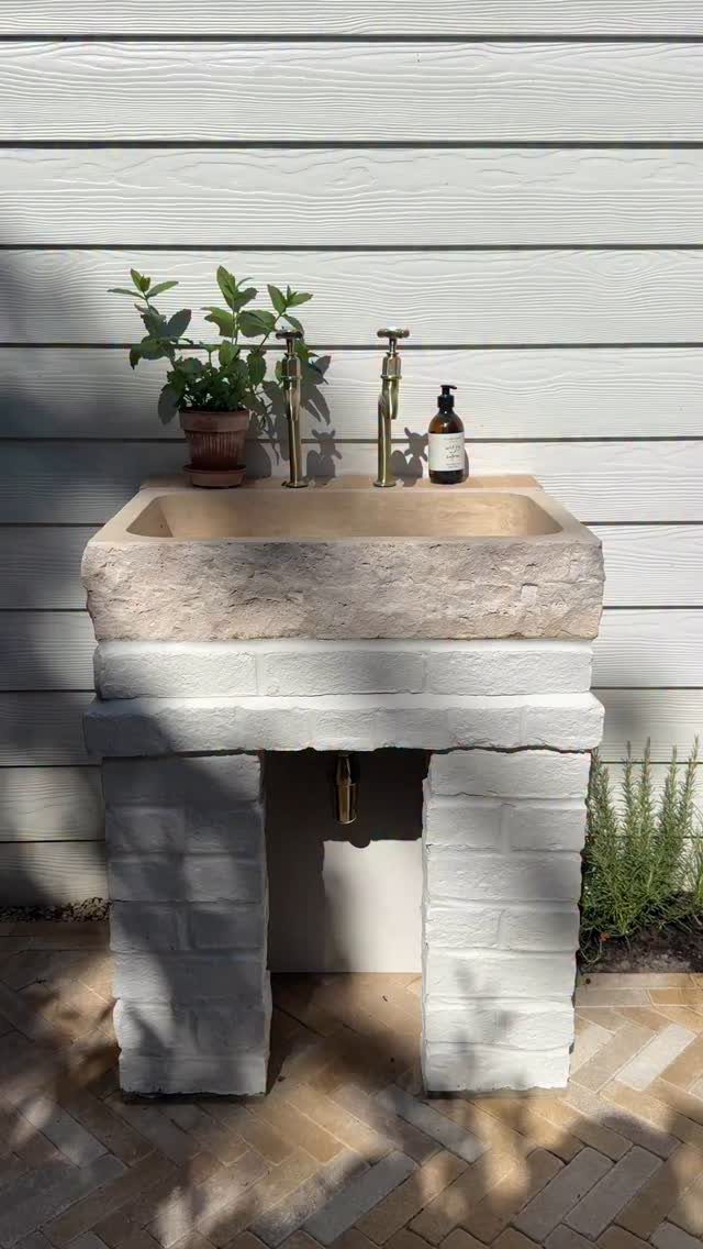 Stone outdoor sink with gold faucets on a white brick base against siding