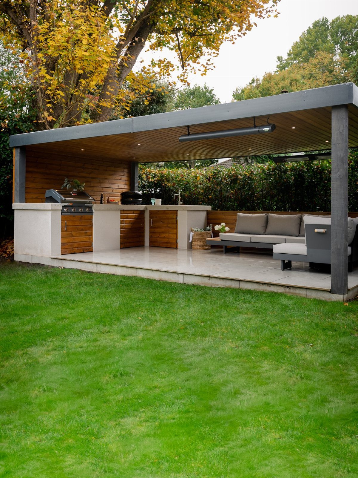 Modern L-shaped outdoor kitchen with white counters and wooden cabinets under a pergola.