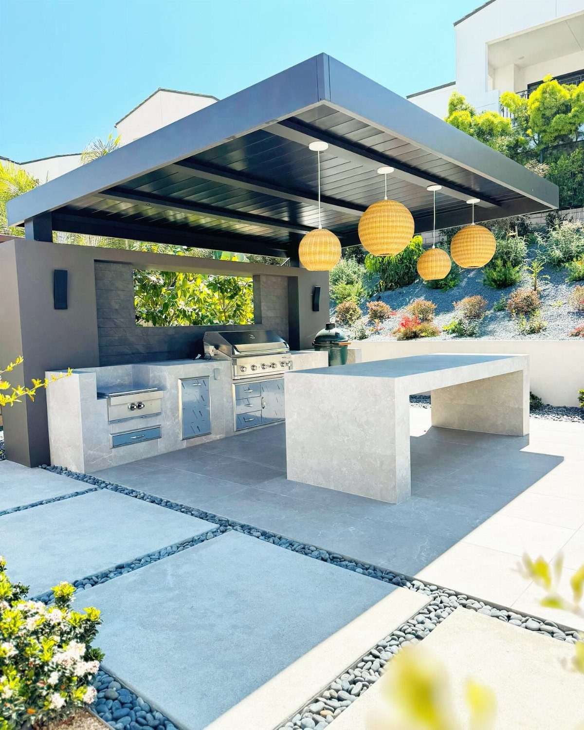 Modern outdoor kitchen with grey concrete surfaces, black pergola, and yellow pendant lights.