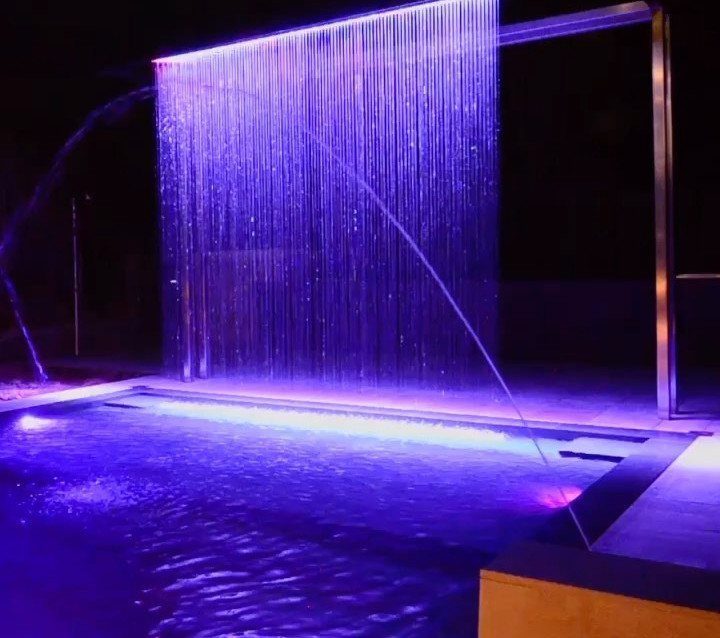 Modern swimming pool at night with a purple illuminated water curtain feature.