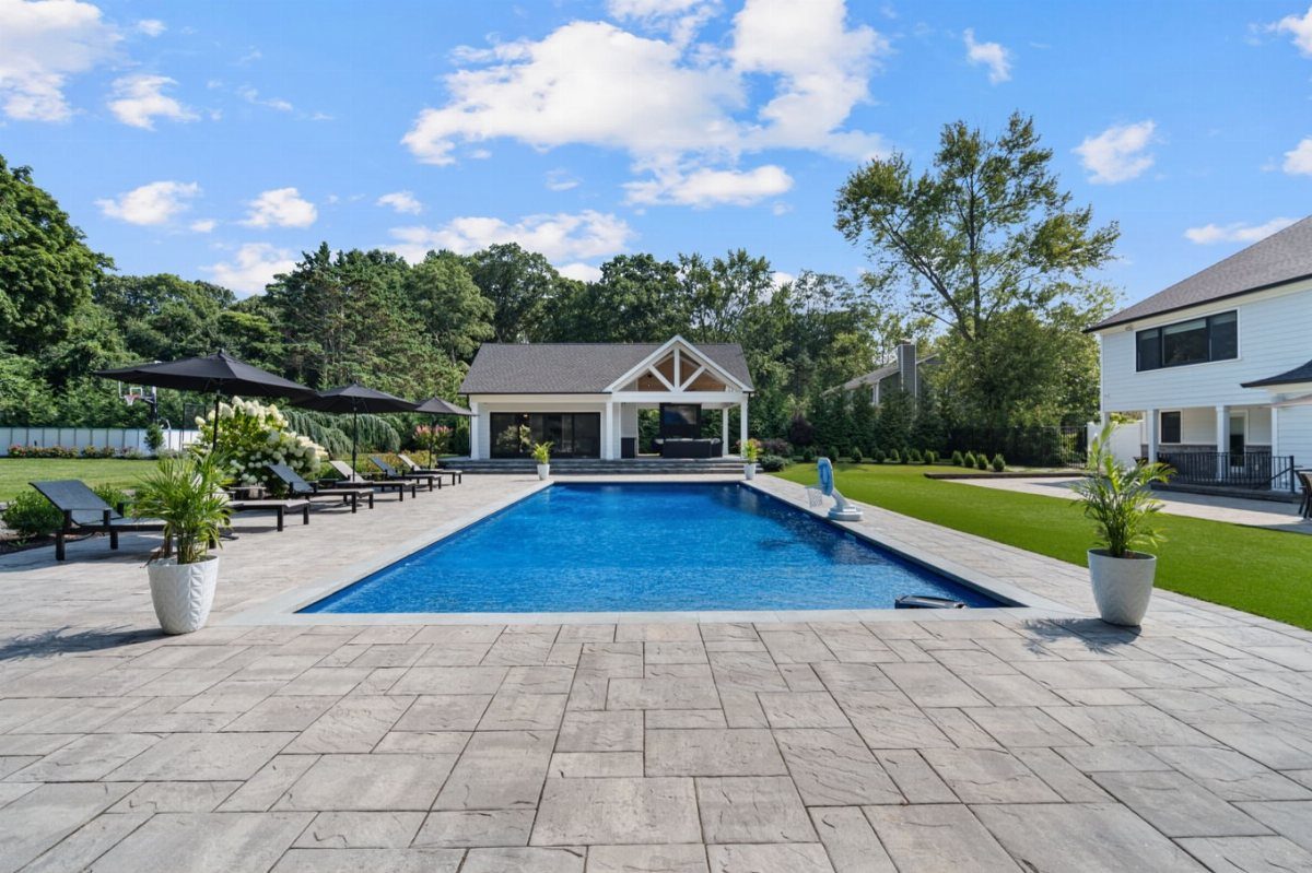 Rectangular blue swimming pool with grey stone patio, black umbrellas, and white pool house.