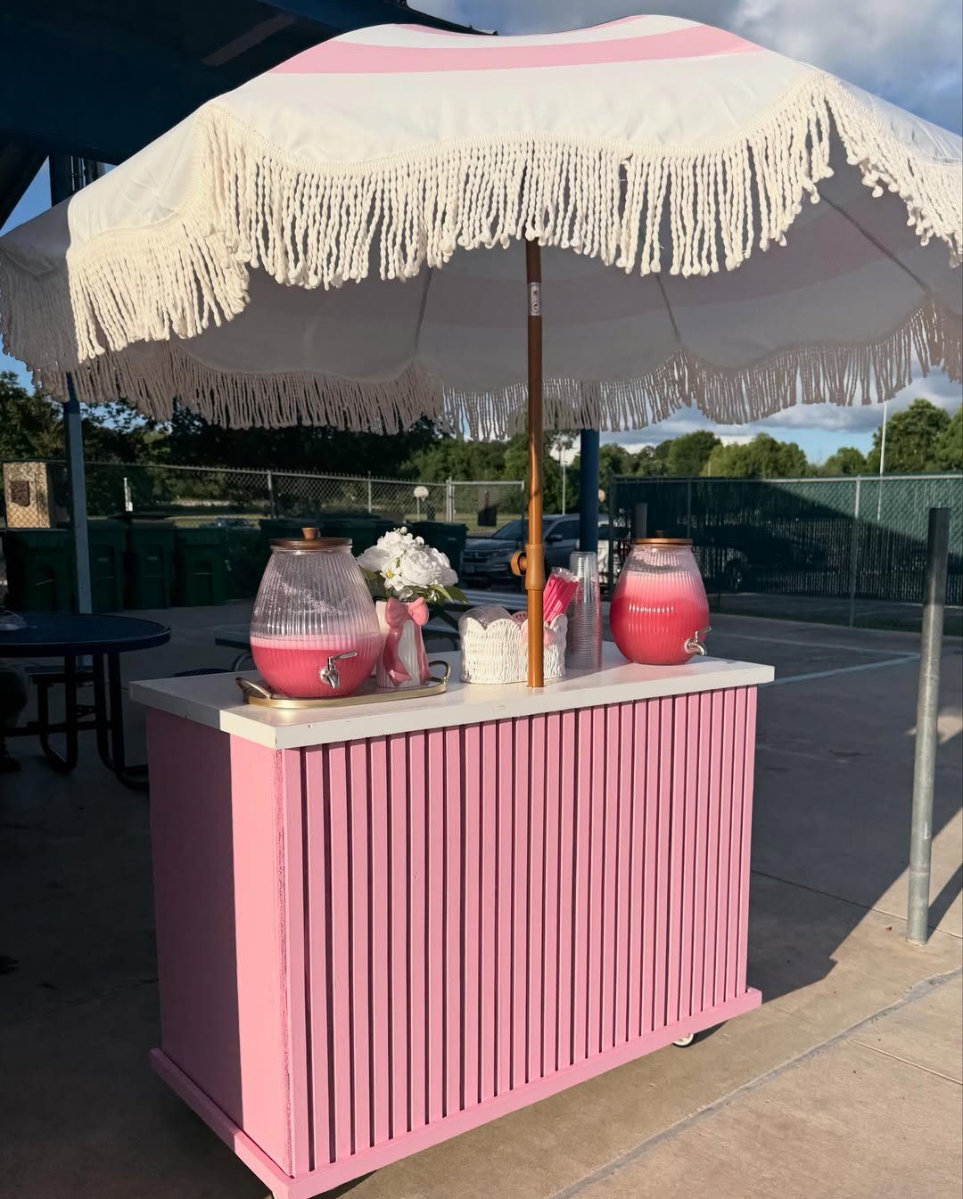 Pink mobile bar cart with beverage dispensers under a white fringed umbrella outdoors.