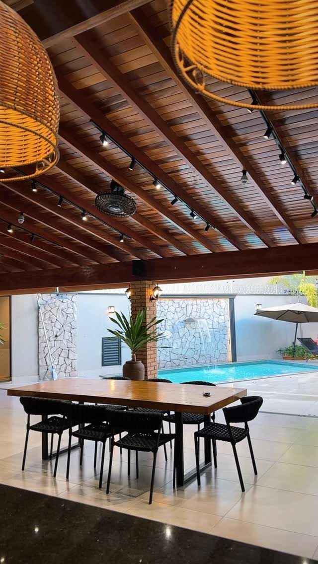 Wooden dining table and black chairs under a patio roof overlooking a swimming pool