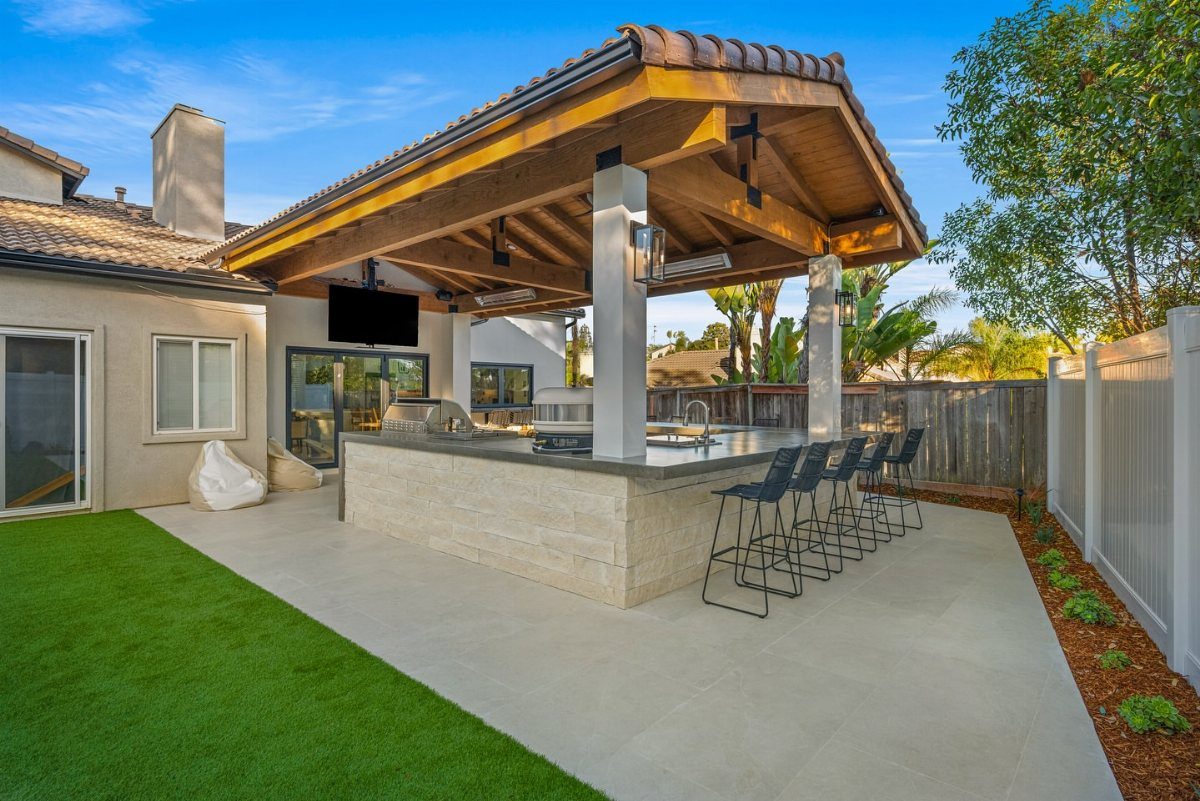 Outdoor kitchen with stone island and bar stools under a wooden pavilion roof.