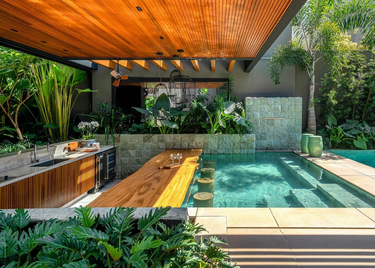 Modern outdoor kitchen and wooden bar beside a turquoise swimming pool with tropical plants.