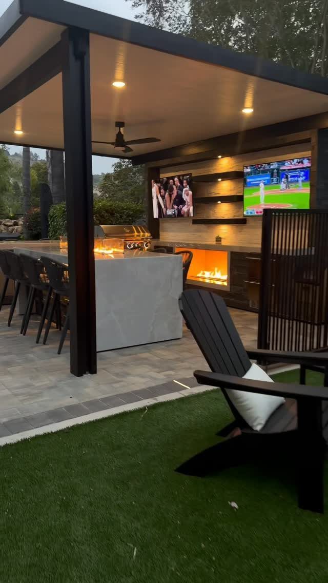 Modern outdoor kitchen with stone island, fireplace, and TVs under a black pergola.