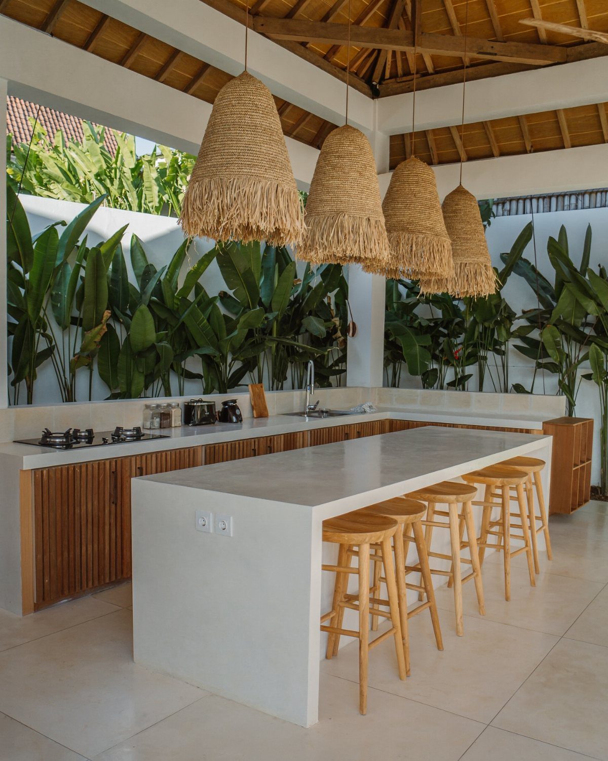 Outdoor kitchen with white concrete island, wooden stools, and straw pendant lights.