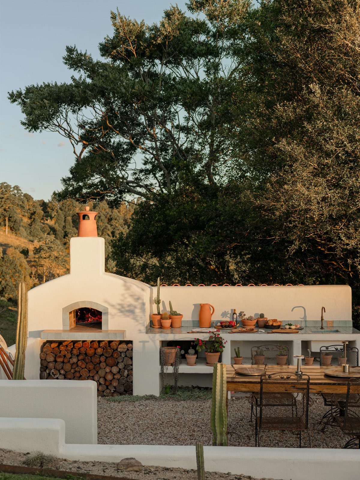White stucco outdoor kitchen with a wood-fired pizza oven and wooden dining table.