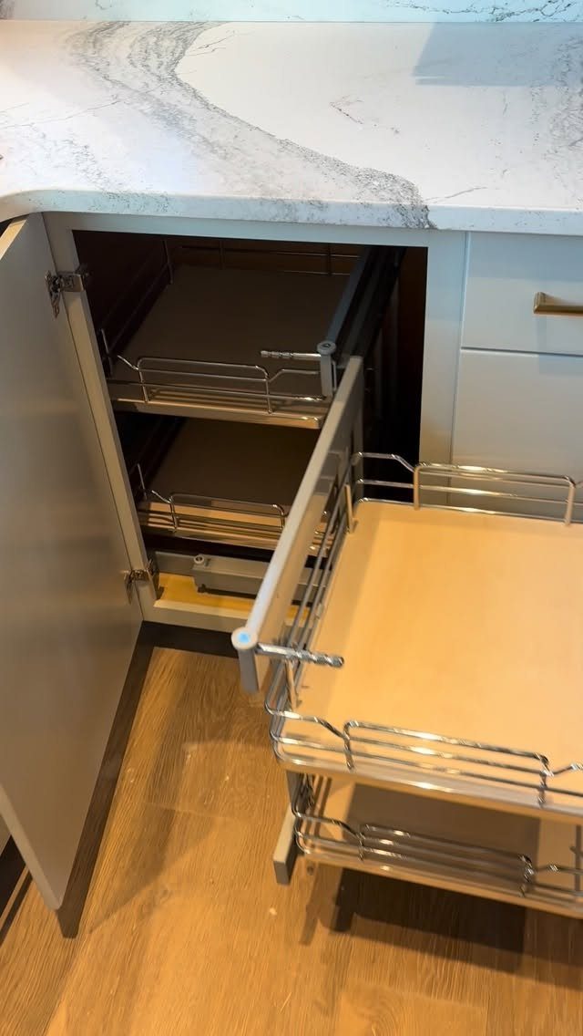 Open grey corner cabinet with pull-out metal wire shelves under marble countertop